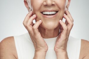 Nose-to-chest closeup of older woman wearing white sweater-vest smiling with her hands on her cheeks